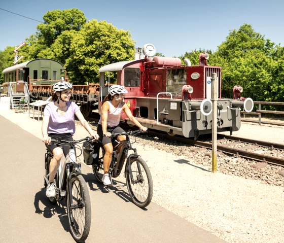 Zwei Radfahrerinnen auf einem Radweg neben einer alten Lokomotive im Eisenbahnmuseum Pronsfeld, umgeben von grünen Bäumen., © Eifel Tourismus GmbH, Dominik Ketz Zwei Radfahrerinnen auf einem Radweg neben einer alten Lokomotive im Eisenbahnmuseum Pronsfeld, umgeben von grünen Bäumen., © Eifel Tourismus GmbH, Dominik Ketz