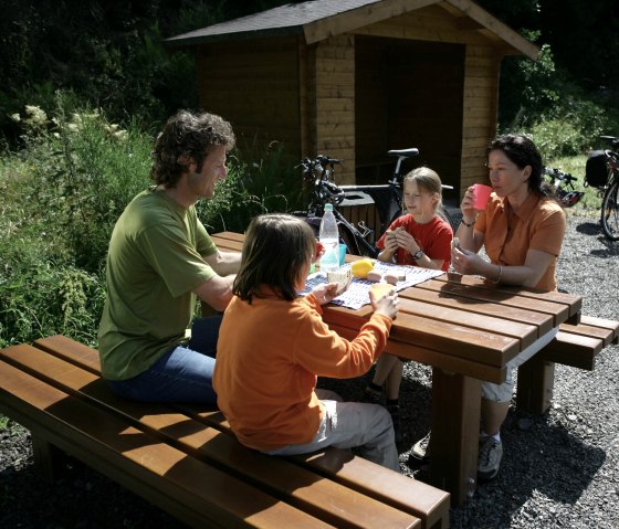 Eine Familie sitzt an einem Holztisch im Freien und macht eine Pause. Im Hintergrund stehen Fahrräder und eine Holzhütte., © Eifel Tourismus GmbH/intention Eine Familie sitzt an einem Holztisch im Freien und macht eine Pause. Im Hintergrund stehen Fahrräder und eine Holzhütte., © Eifel Tourismus GmbH/intention