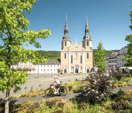 Ein Radfahrer fährt vor der Basilika in Prüm entlang, umgeben von grünen Bäumen und bunten Pflanzen, bei klarem Himmel., © Eifel Tourismus GmbH, Dominik Ketz Ein Radfahrer fährt vor der Basilika in Prüm entlang, umgeben von grünen Bäumen und bunten Pflanzen, bei klarem Himmel., © Eifel Tourismus GmbH, Dominik Ketz