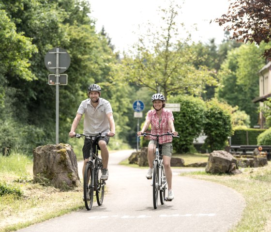 Zwei Personen fahren mit Fahrrädern auf einem Radweg bei Plein. Sie tragen Helme und sind von grüner Natur umgeben., © Eifel Tourismus GmbH, D. Ketz Zwei Personen fahren mit Fahrrädern auf einem Radweg bei Plein. Sie tragen Helme und sind von grüner Natur umgeben., © Eifel Tourismus GmbH, D. Ketz