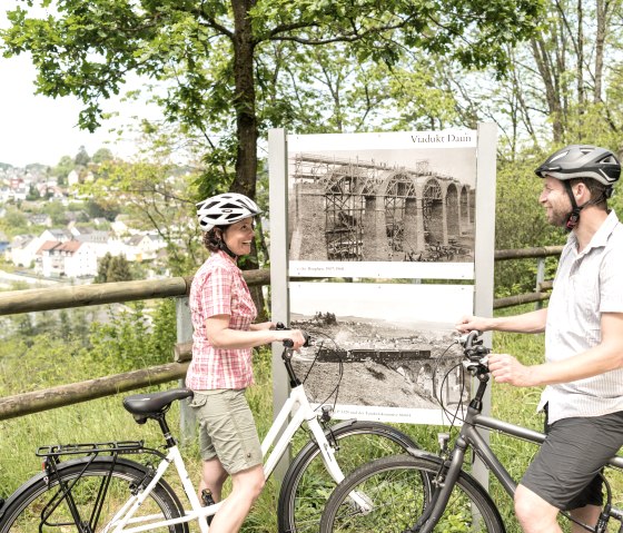 Zwei Radfahrer betrachten historische Fotos des Viadukts Daun am Maare-Mosel-Radweg. Im Hintergrund ist eine Stadtlandschaft zu sehen., © Eifel Tourismus GmbH, D. Ketz Zwei Radfahrer betrachten historische Fotos des Viadukts Daun am Maare-Mosel-Radweg. Im Hintergrund ist eine Stadtlandschaft zu sehen., © Eifel Tourismus GmbH, D. Ketz