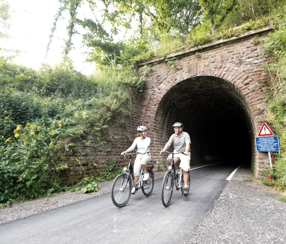Two cyclists ride out of an overgrown tunnel on the Vennbahn near Lommersweiler. There is a warning sign at the edge., © vennbahn.eu Two cyclists ride out of an overgrown tunnel on the Vennbahn near Lommersweiler. There is a warning sign at the edge., © vennbahn.eu