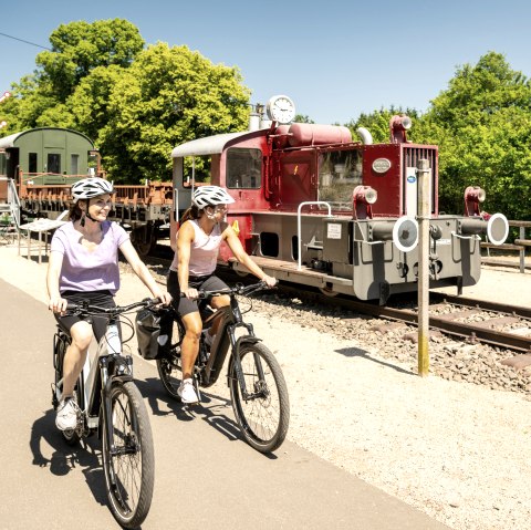 Zwei Radfahrerinnen auf einem Radweg neben einer alten Lokomotive im Eisenbahnmuseum Pronsfeld, umgeben von grünen Bäumen., © Eifel Tourismus GmbH, Dominik Ketz Zwei Radfahrerinnen auf einem Radweg neben einer alten Lokomotive im Eisenbahnmuseum Pronsfeld, umgeben von grünen Bäumen., © Eifel Tourismus GmbH, Dominik Ketz