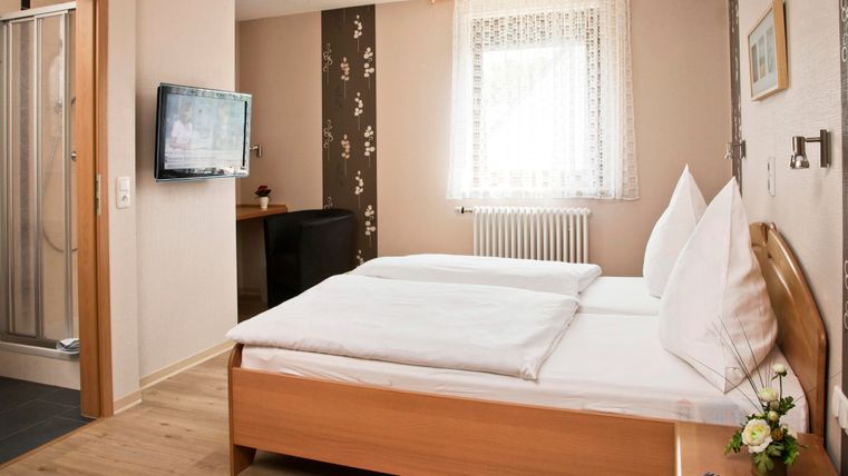 Ein helles Zimmer mit zwei Betten und einem Fernseher. Das Badezimmer ist durch eine Glaswand abgetrennt und es gibt eine schöne Fensterfront.