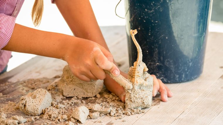 Un enfant façonne du sable avec une main, tandis qu'il tient une petite figurine. En arrière-plan, il y a un seau bleu.
