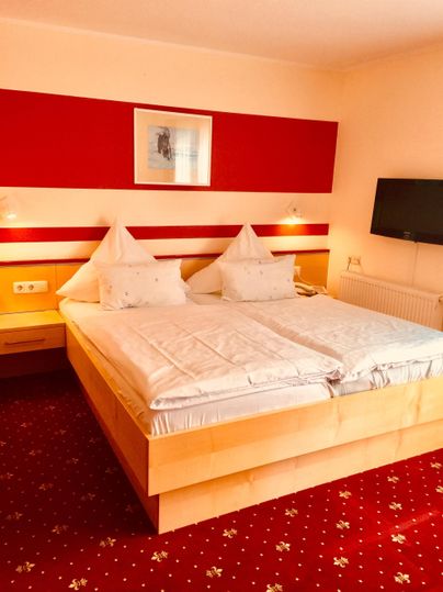 Ein gemütliches Schlafzimmer mit einem Doppelbett und roten Akzenten. Es gibt einen Fernseher an der Wand und ein schönes Teppichmuster auf dem Boden.