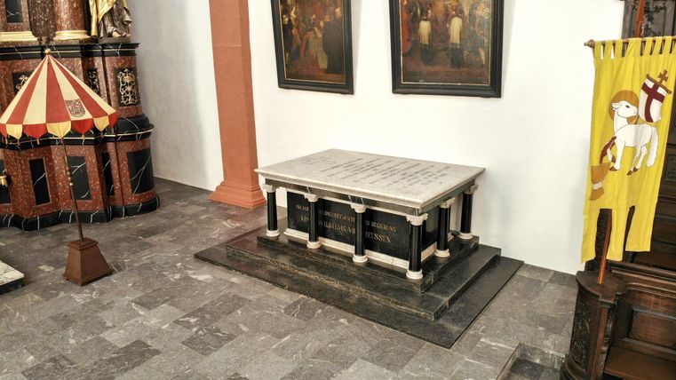 Ein Raum mit einem Steinsarg auf einer Plattform. Im Hintergrund sind mehrere Bilder und ein Fahne zu sehen.