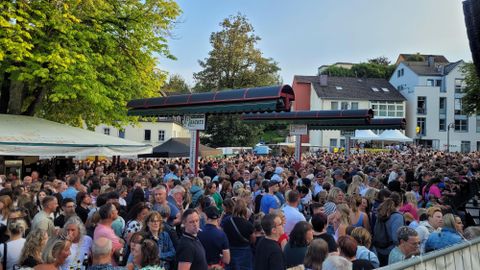 Eine große Menschenmenge versammelt sich an einem Veranstaltungsort. Im Hintergrund sind einige Gebäude und Bäume sichtbar.