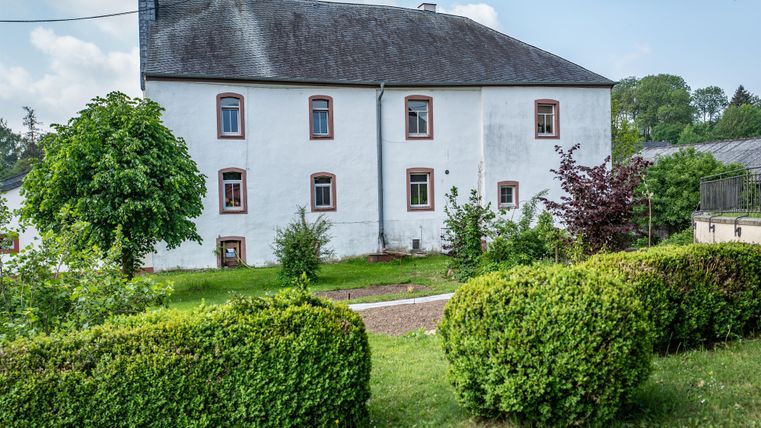 Ein altes, weißes Haus umgeben von einem grünen Garten. Bäume und Sträucher verleihen der Szene eine ruhige Atmosphäre.