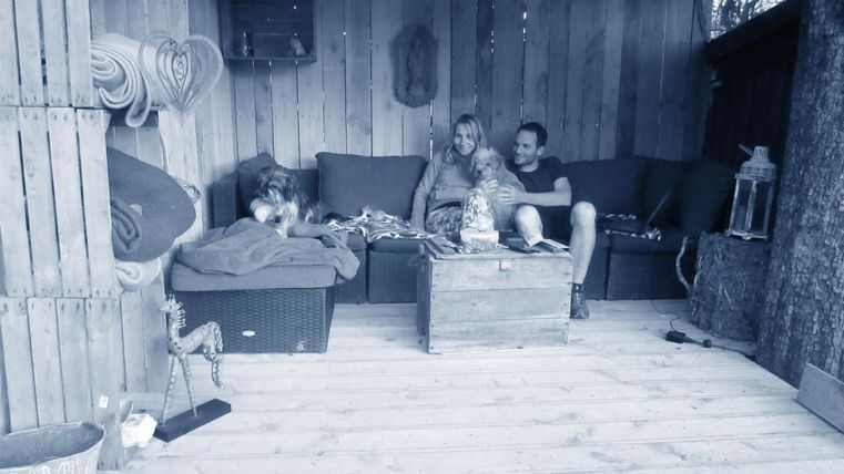 Ein gemütlicher Raum mit Holzwänden und einer Sitzgelegenheit. Zwei Personen und zwei Hunde entspannen sich auf einem Sofa.