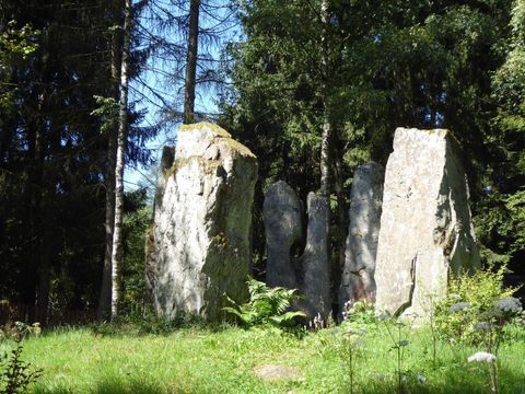 A group of large, upright stones stands in a wooded setting. Surrounded by grass and trees, the scene radiates a mystical atmosphere.