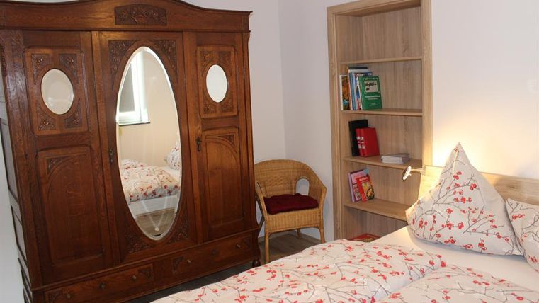 Ein gemütliches Schlafzimmer mit einem antiken Kleiderschrank und einem Bücherregal. Das Bett ist mit blütenbedruckten Kissen dekoriert.