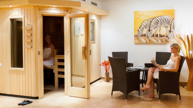 Ein modernes Spa mit einer Holzsauna auf der linken Seite. Rechts sitzt eine Frau in Bademode und liest entspannt an einem Tisch.