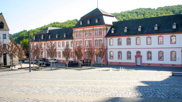 Ein historisches Gebäude mit rotem und weißem Mauerwerk steht an einem Platz. Im Hintergrund sind grüne Hügel und ein klarer Himmel zu sehen.