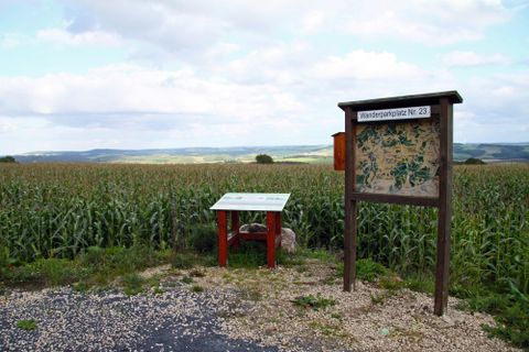 Ein Informationsstand in einem Feld mit Mais. Im Hintergrund sind sanfte Hügel und ein bewölkter Himmel zu sehen.