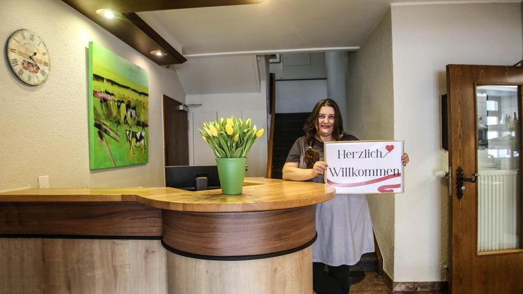 Eine freundliche Rezeption mit einer lächelnden Mitarbeiterin. Sie hält ein Schild mit der Aufschrift „Herzlich Willkommen“ und es stehen frische Blumen auf dem Empfangstresen.