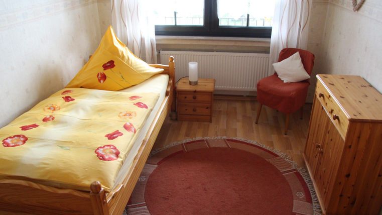 Ein gemütliches Zimmer mit einem Einzelbett, einem kleinen Tisch und einem Stuhl. Es gibt einen runden Teppich und viel Tageslicht durch das Fenster.