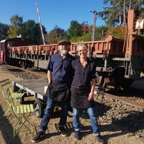 Die beiden Betreiber des Café Stullwerk stehen vor den Bahngleisen und der Lok am Café und lächeln uns bei Sonne an. 