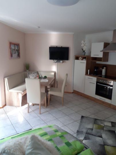 Ein gemütlicher Wohnraum mit einer kleinen Essecke und einer modernen Küche. Die Wände sind hell gestrichen und es gibt ein Fenster, das Licht hereinflutet.