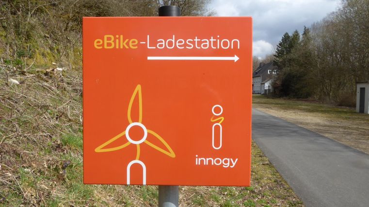 Ein orangefarbener Hinweis auf eine eBike-Ladestation mit einem Windkraftsymbol. Die Ladestation befindet sich in Richtung des angezeigten Pfeils.