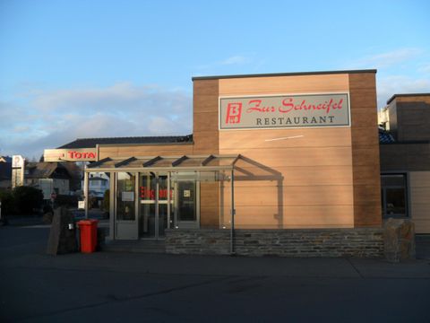 Ein modernes Restaurant mit der Aufschrift „Feuer Schnitzel“. Die Fassade ist aus Holz und Glas und es gibt einen Eingangsbereich mit Überdachung.