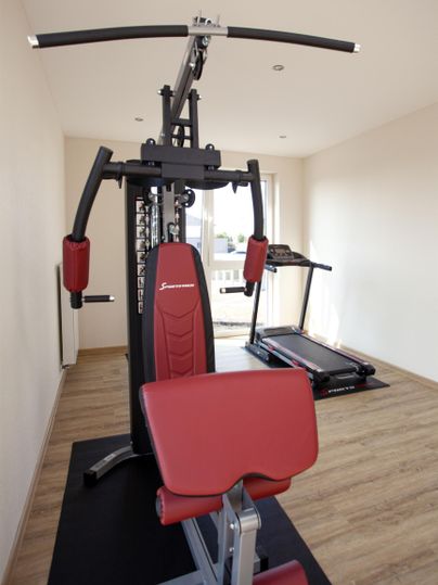 Ein modernes Fitnessstudio mit einer Kraftstation und einem Laufband. Der Boden ist mit Holz ausgelegt und der Raum ist gut beleuchtet.