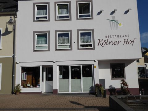 Ein Restaurant namens "Kölner Hof" mit einer hellen Fassade und großen Fenstern. Die Umgebung ist gepflastert und es gibt einige Pflanzen vor dem Eingang.