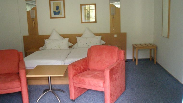 Ein einfaches Zimmer mit zwei roten Sesseln und einem Tisch. Im Hintergrund sieht man ein Doppelbett mit weißen Kissen.