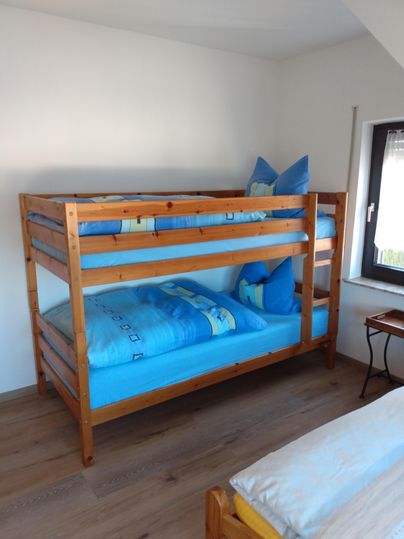 Ein Kinderbett mit zwei Etagen aus Holz steht in einem hellen Zimmer. Die Bettwäsche ist blau und es gibt bunte Kissen.