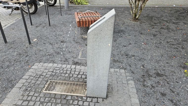 Ein grauer Stein mit einer schrägen Oberfläche steht in einem gepflasterten Bereich. Im Hintergrund sind ein paar Bänke und Bäume sichtbar.