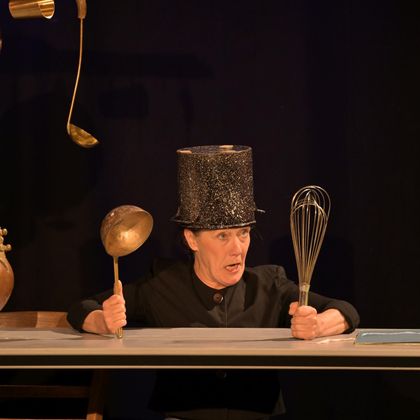 Une personne avec un chapeau haut de forme scintillant tient une cuillère en bois et un fouet. Elle se tient derrière une table et a une expression marquée.