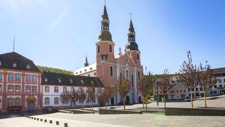 Eine beeindruckende Kirche mit zwei Türmen und einer schönen Fassade in einer ruhigen Stadt. Im Vordergrund sind gepflanzte Bäume und ein offener Platz zu sehen.
