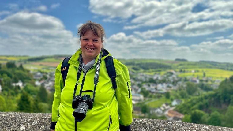 Eine Frau mit einer Kamera steht an einer Aussichtsstelle. Im Hintergrund sind grüne Hügel und ein kleines Dorf zu sehen.