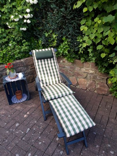 Ein bequemer Liegestuhl mit kariertem Polster steht vor einer grünen Hecke. Neben dem Stuhl steht ein kleiner Tisch mit einer Pflanze.