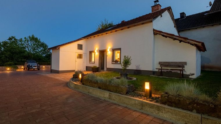 Ein gemütliches Haus mit weißer Fassade und einem roten Dach. Der Eingangsbereich ist gut beleuchtet und von einem gepflasterten Weg umgeben.