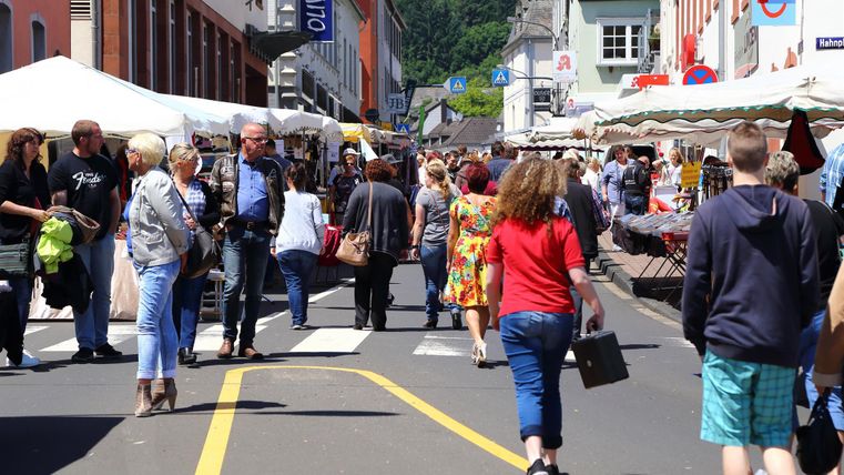 Ein belebter Markttag auf einer Straße, umgeben von Ständen und vielen Menschen. Die Atmosphäre ist lebhaft und freundlich, mit bunten Kleidungsstücken und eingekauften Waren.