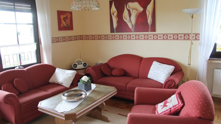 Ein gemütliches Wohnzimmer mit roten Sofas und einem Holztisch. An den Wänden hängen dekorative Bilder und es gibt viel Tageslicht.