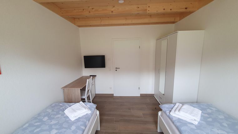 Ein schlichtes Zimmer mit zwei Einzelbetten und einem Holzdecke. Im Raum befinden sich ein Fernseher, ein Schreibtisch und ein Kleiderschrank.
