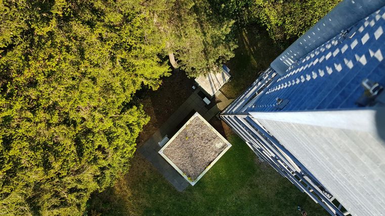 Eine Vogelperspektive auf einen Garten mit einem kleinen Pavillon und umgebenden Bäumen. Im Hintergrund ist ein Hochhaus zu sehen.