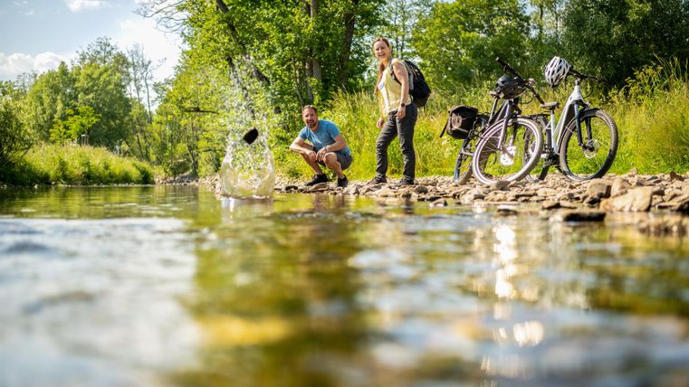 Two people are standing by the bank of a river, throwing stones into the water. Next to them are two bicycles. The surroundings are green and sunny.