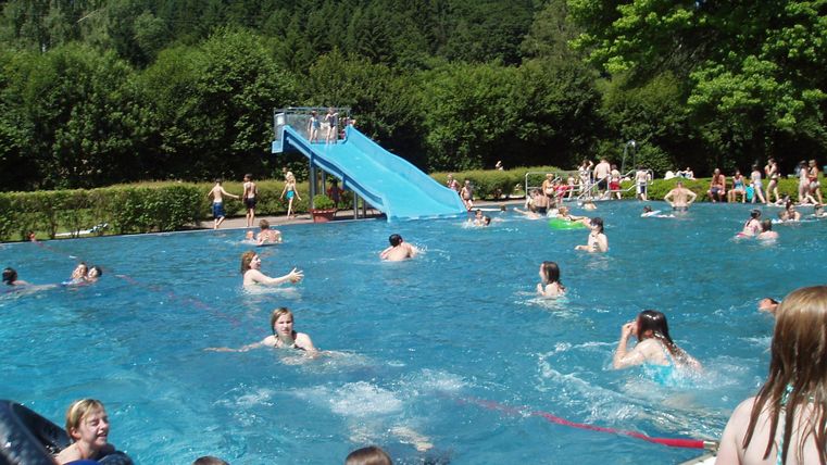 Ein lebhafter Schwimmbecken mit vielen Menschen, die sich im Wasser vergnügen. Im Hintergrund ist eine große Rutsche zu sehen.
