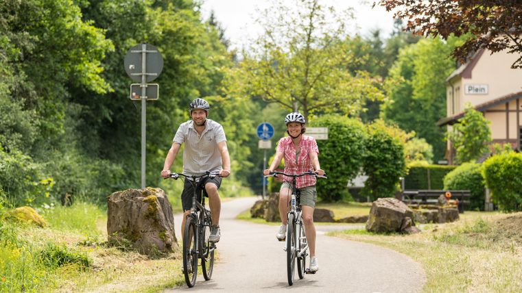 Zwei Radfahrer auf einem Radweg bei Plein, umgeben von grüner Natur.