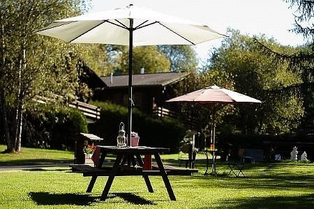 Ein schöner Garten mit einem Holztisch unter einem Sonnenschirm. Im Hintergrund sind weitere Sonnenschirme und grüne Bäume zu sehen.