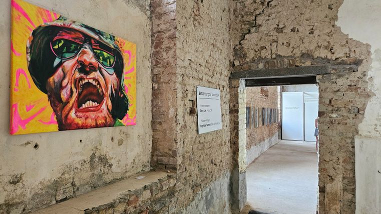 Ein buntes Gemälde eines Gesichts hängt an einer gemauerten Wand in einem alten Raum. Durch einen offenen Durchgang sieht man einen weiteren Raum im Hintergrund.