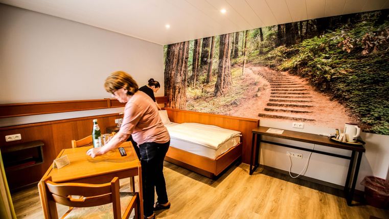 Ein gemütliches Zimmer mit einem Tisch und einem Stuhl. An der Wand ist ein großes Bild eines Waldpfades zu sehen.