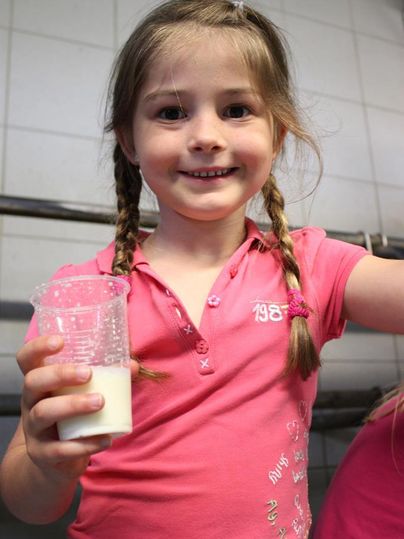 Ein lächelndes Mädchen mit zwei Zöpfen hält ein Glas Milch in der Hand. Sie trägt ein pinkes T-Shirt.