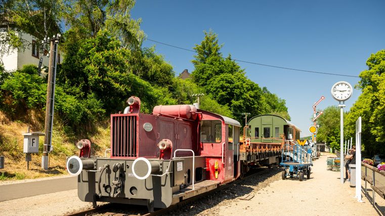 Une locomotive rouge est arrêtée sur une voie avec des wagons. À l'arrière-plan, des arbres et un panneau sont visibles.