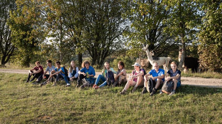 Eine Gruppe von Menschen sitzt auf einer Wiese. Im Hintergrund sind einige Bäume und Lamas zu sehen.