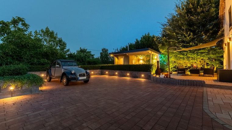 Eine moderne Einfahrt mit einem kleinen Auto und einer gemütlichen Terrasse. Die Umgebung ist gut beleuchtet und von Bäumen umgeben.