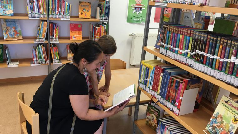 Une mère et son enfant sont assis dans une bibliothèque et regardent ensemble un livre. En arrière-plan, on peut voir des étagères avec de nombreux livres colorés.